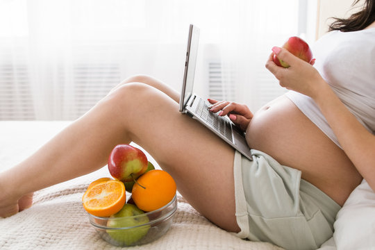 Pregnant woman eats apple while chatting in laptop. Work and relax combining, fresh fruits and healthcare concept of special mother-to-be period