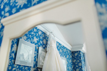 Mirror's reflection of cream bridal dress which hangs on the cornice on a background the blue floral pattern wall.