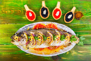 Fish trout cooked grill in foil with lemons, tomatoes and greens. Next  tartlets with red caviar in the form of spoons. On green wooden  background. Top viwe.