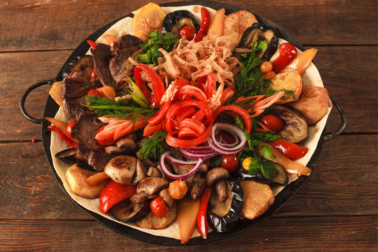 Dish with stewed meat, different kind of vegetables, field mushrooms, laid on lavash and big copper plate, and decorated with sliced onion and pepper. Top view picture on wooden table