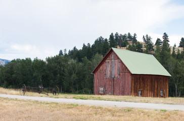 Obraz premium Old red barn near treed ridge, with rustic farm equipment.