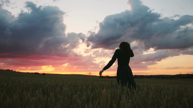 Young beatiful woman in black dress spinning and having fun in the green field on the colorful sky background during the sunset.