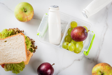 Back to school. A healthy lunch in a box is fresh fruit (apples, plums, grapes), a bottle of yogurt and a sandwich with lettuce, tomatoes, cheese, meat. White marble table. Copy space top view
