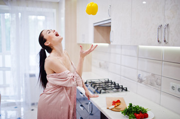 Portrait of a beautiful young woman in pink robe posing with a yellow pepper in her kitchen.