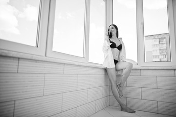 Portrait of a lovely girl in underwear and male shirt standing with a glass of water in her hands on the balcony. Black and white photo.