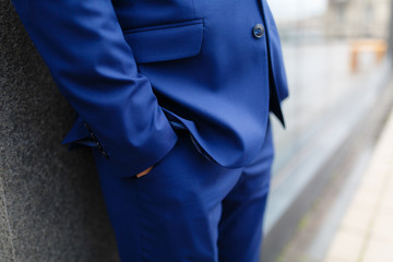 A man in a blue shiny suit holds his hands in his pockets standing at the stone wall. close up of businessman hand in his pockets with wedding ring