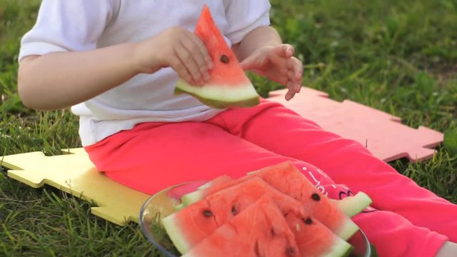 A little baby girl sits on a green grass on a multi-colored rug and takes a piece of a juicy watermelon from a glass cup, then bites it on the background of a pond in the summer at sunset