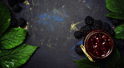 Blackberry jam, fresh berries and green leaves on brown background, top view
