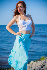 Redhead slim woman wearing tender pastel dress posing on a sea, fashionable woman walking on the shore. Young girl on a beach in a white tones. Pastel colors. The concept of femininity ad harmony