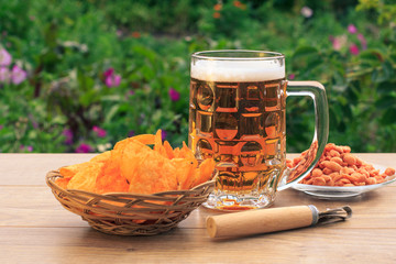 Glass mug of beer on wooden table with potato chips in wicker basket, peanuts in plate and opener