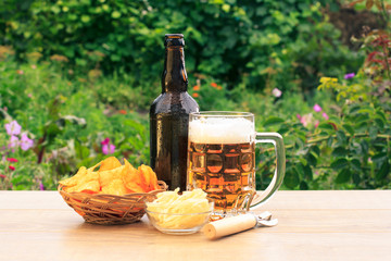 Glass mug of beer and bottle of beer on wooden table with potato chips in wicker basket, dried...