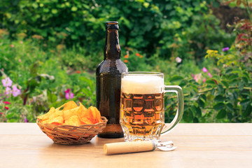 Glass mug of beer and bottle of beer on wooden table with potato chips in wicker basket and opener