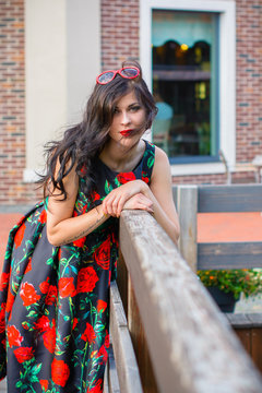 Gorgeous vintage brunette woman with curly hair on a style 50s in black dress with red roses. Pretty girl walk on a streets and have a good mood feeling wonderful