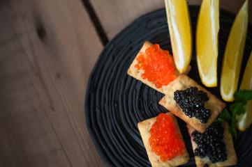 Caviar on the cracker. Black and red caviar on a bread slice on a black plate. Caviar, lemons, parsley in the dark on the background of a wooden table