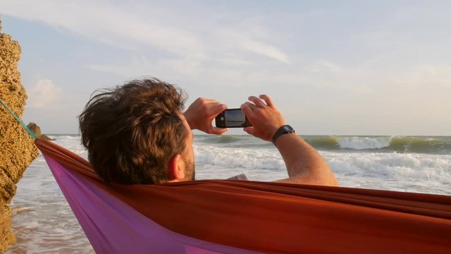 Young Man Relaxing in Hammock on a Beach and Taking Photo of Sea with Mobile Phone. HD Slowmotion. Crimea, Russia.