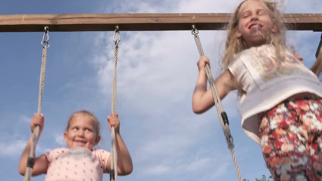 Happy children on wood swing set against blue sky on a sunny day. Two cute little girls have fun together swinging high on playground in backyard