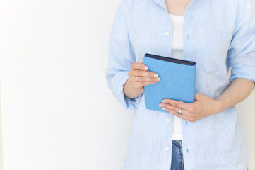 Young Girl in blue cotton shirt standing near white wall holding blue notebook in hands