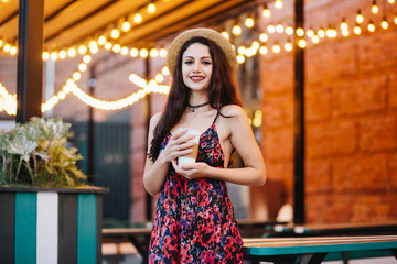 Smiling young woman with black long hair and make-up, dressed in summer dress and hat, holding fresh coffee going to drink it at terrace. Beautiful female having drink at cozy cafe or restaurant