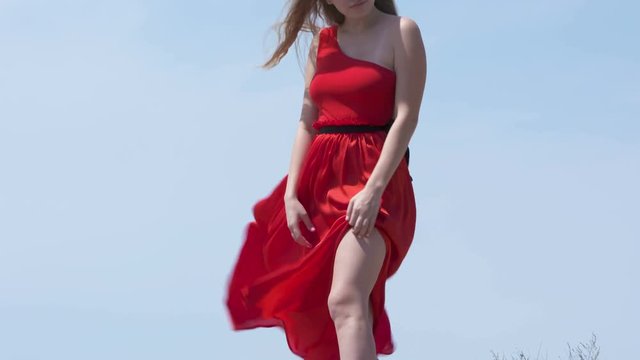Young barefoot girl in long red dress on an old stone wall against blue sky. Red dress blowing in the wind in slow motion.