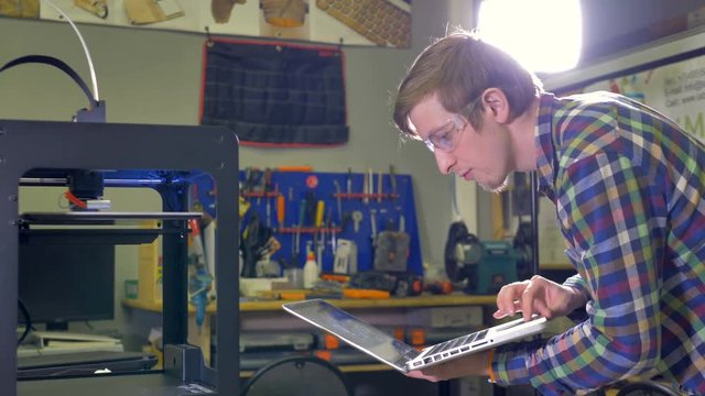 A man uses a laptop touchpad to set a 3d-printer. 
