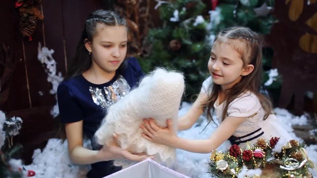 Two little girls at a Christmas tree prepare a gift for the grandmother. Granddaughters pack a white shawl as a present to their beloved grandmother in a gift box.