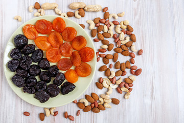 On a light table, a plate of dried dried apricots and prunes. Near it are scattered nuts. A mixture of nuts consists of peeled and peanuts, almonds and cashews.