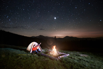 Night camping in the mountains. Couple tourists sitting in the illuminated tent near campfire, looking at night starry sky. Man pointing at the sky. Long exposure