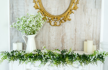 White decorative fireplace with candles on it near wooden wall. Floral decoration of white flowers and greenery over white fireplace