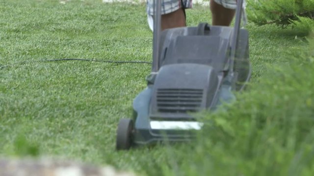 Lawn Mowing In The Garden. Man In Shorts And Flip Flops Cutting Grass In His Yard With Corded Electric Lawn Mower.