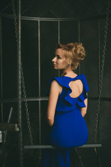 Красивая девушка в вечернем синем платье сидит на качелях в клетке в интерьерной студии