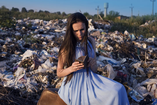 A Girl Among A Garbage Dump Looking Into An Empty Purse With Sadness