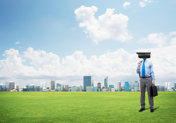 Camera headed man standing on green grass against modern cityscape