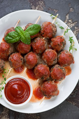Top view of baked meatballs on skewers served with tomato sauce on a white plate, vertical shot