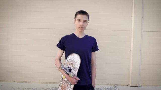 Image of handsome teenager standing in the street and holding skateboard in his hands. Boy spending free time in skateboarding park demonstrating healthy lifestyle and looking at the camera.
