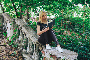 Young beautiful blonde military style dressed student sitting on the steps of the stairs in the old Park and reads a book