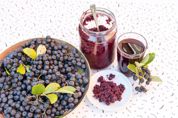 Fresh juice and jam of black chokeberry (Aronia melanocarpa) in glass and berry in pot on white textured background