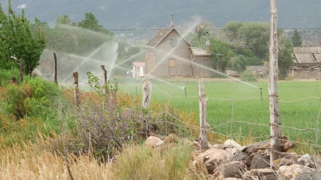 An old wooden cabin sits in a mountain valley, behind a wooden fence as sprinklers irrigate a surrounding field.