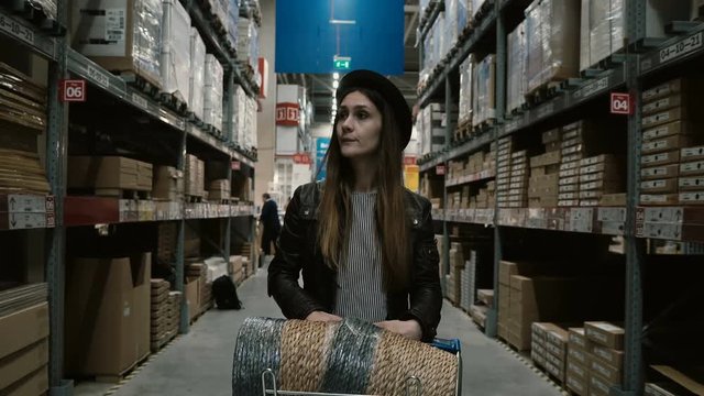 Young beautiful woman with cart full of goods walking through the high shelves with things for repair in big warehouse.