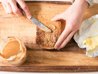 High angle view of woman's hands spreading peanut butter on wholemeal toast on wooden board (selective focus)