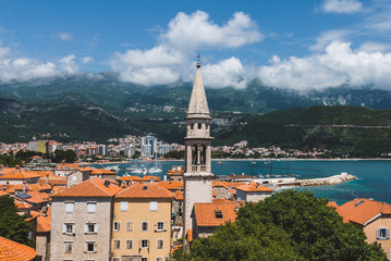 Naklejka premium Budva Old town view from above to city roofs, St.John church spire, cloudy mountains and Adriatic sea on background. Tourist capital Budva high view, Montenegro.