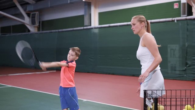 Image of lesson at indoor court with professional equipment. Child learning process of correct using a racket. Boy playing tennis with female coach in white outfit giving him yellow balls.