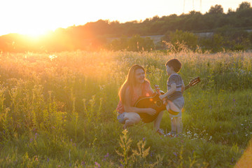 Woman play guitar with a little boy in nature. Mom and her son playing guitar on meadow in sunset