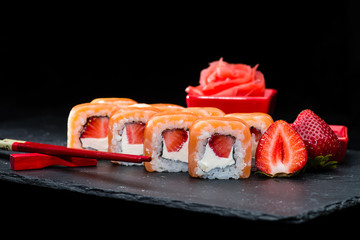 Traditional Japanese cuisine. Selective focus on sweet sushi rolls with salmon, cream cheese, rice and strawberry on dark background, near strawberries and ginger