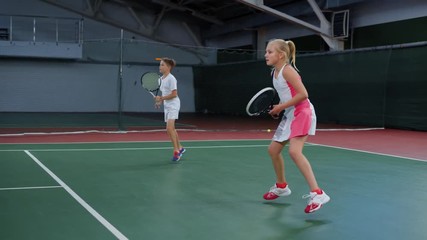 Two young athletes in recreation area playing sport game. Happy sister and brother having tennis lesson spending time at indoor court. Girl and boy serving and returning yellow balls with rackets.