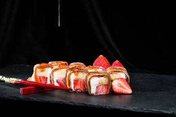 new trend in the Japanese cuisiune. Sweet sushi rolls in pancake with cream cheese, strawberry and banana throwing with jam, dark background
