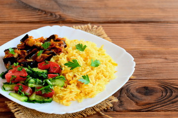 Simple veggie risotto, tomato and cucumber salad, stewed eggplant on a white plate. Brown wooden background with copy space. Summer risotto from rice and vegetables. Vegan menu