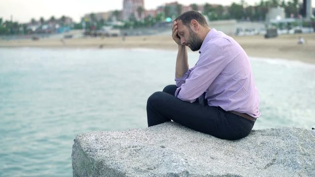 Sad, unhappy businessman sitting on rock near sea
