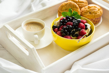 Wooden tray with tasty breakfast on bed. Espresso, banana muffins, cottage cheese with blueberry and raspberry. Sun light. Selective focus
