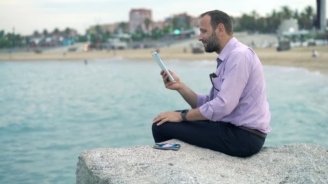 Young businessman using tablet computer sitting on rock near sea 
