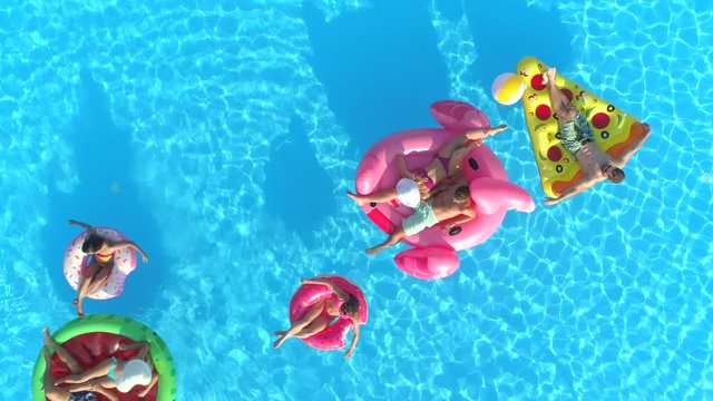 AERIAL TOP DOWN Relaxed girls and guys laying on colourful floaties floating on pool water. Happy smiling friends enjoying summer vacation on inflatable watermelon, pizza, flamingo and doughnut floats